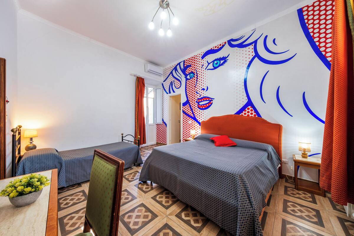 A hotel room with two beds, a large wall mural, patterned flooring, and a small table with a lamp.
