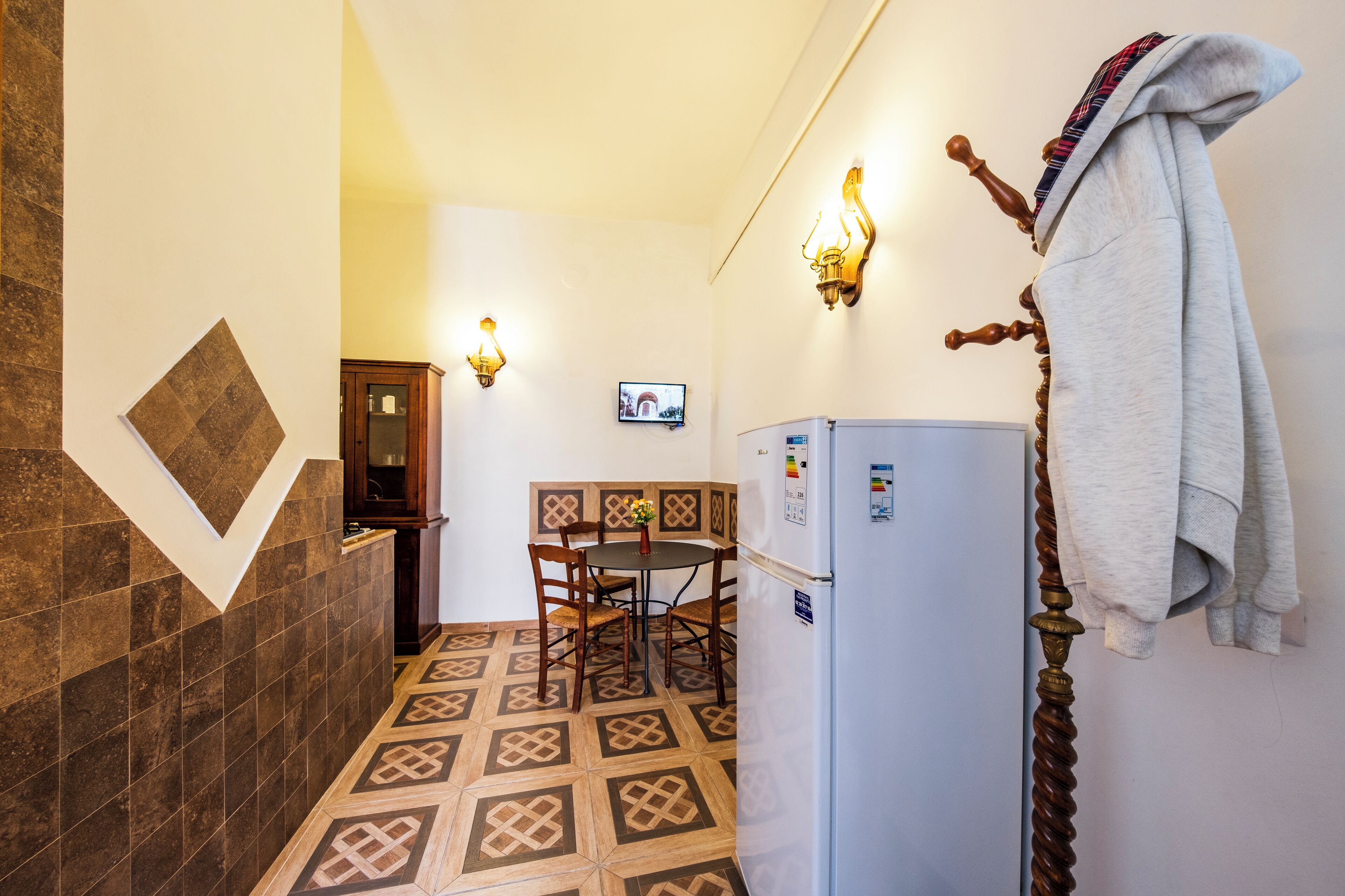 A small kitchen with a tiled floor, a refrigerator, a dining table with chairs, and a television mounted on the wall.