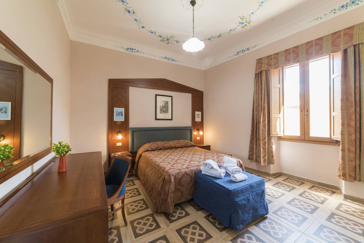 A hotel room with a bed, a desk, a chair, a small table with towels, a potted plant, and a window with curtains.