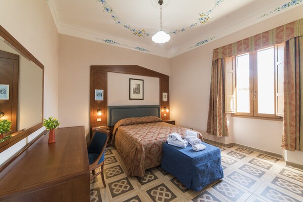 A hotel room with a bed, a desk, a chair, a small table with towels, a potted plant, and a window with curtains. - Affittacamere Al Seminario (Tivoli)