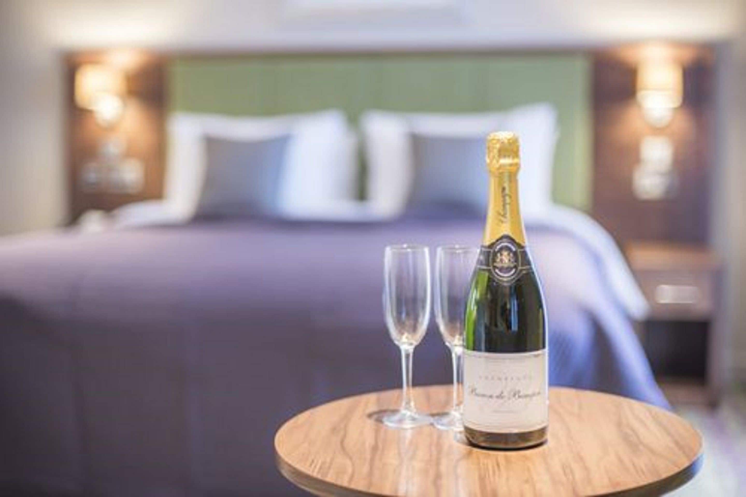 A bottle of champagne on a wooden table with two glasses, in a hotel room with a bed and nightstands in the background.
