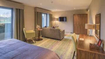 Habitación de hotel con una cama grande, un sofá, un escritorio con lámpara, un televisor y vistas a un jardín por la ventana.