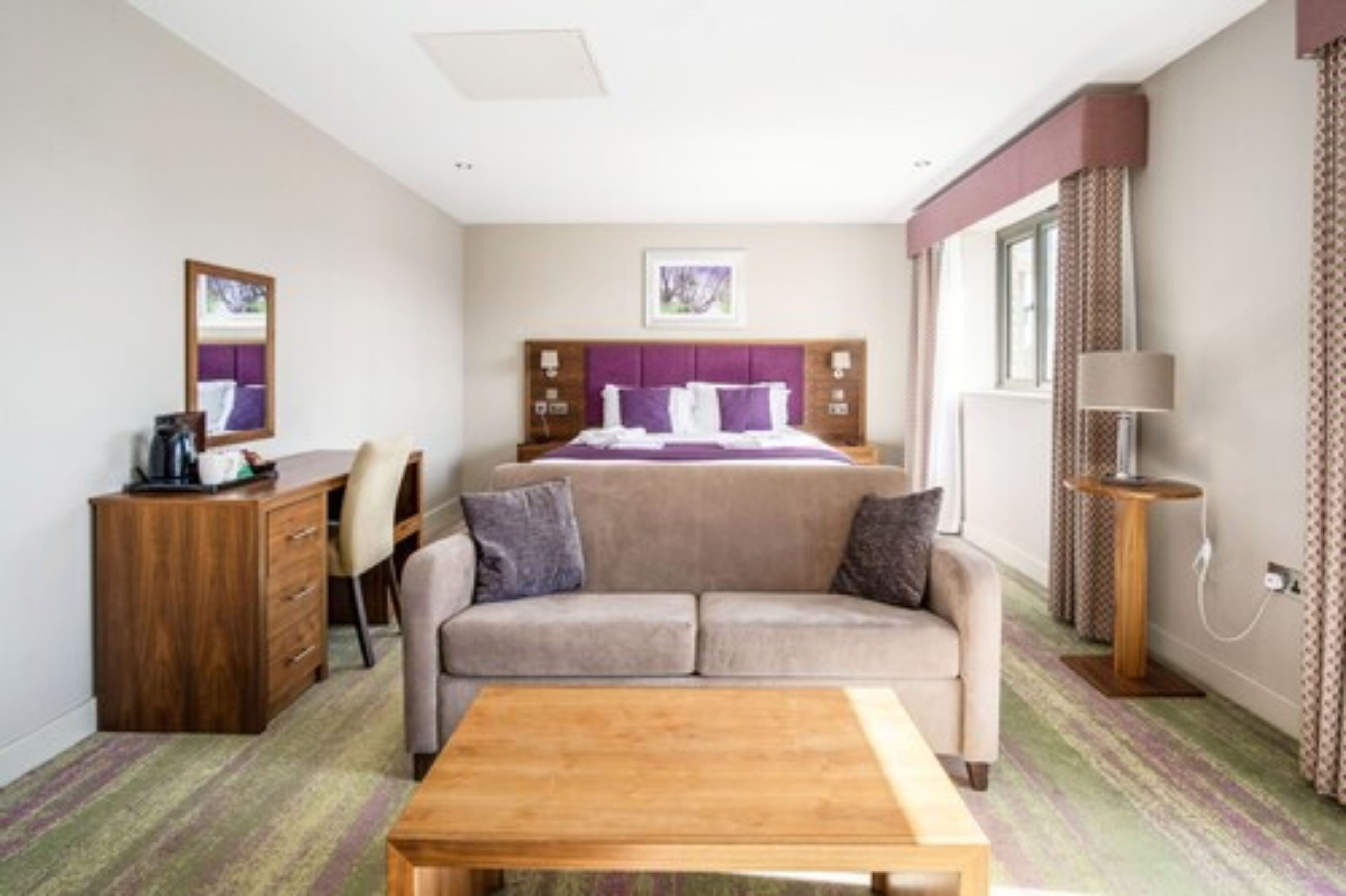 A hotel room with a bed, a sofa, a wooden table, a desk, and a lamp.
