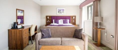 A hotel room with a bed, a sofa, a wooden table, a desk, and a lamp.