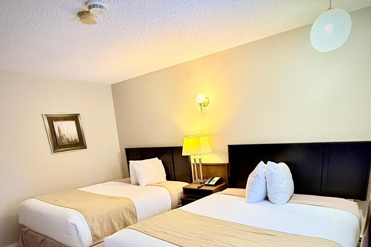 A hotel room with two beds, a nightstand with a lamp, a framed picture on the wall, and a ceiling light fixture.