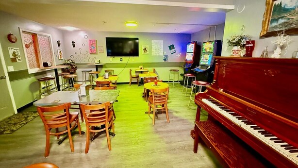A room with a piano, tables, chairs, a TV, and arcade games. - Melville Country Inn (Melville)