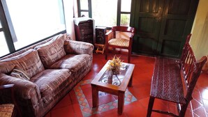 Un salon avec un canapé à motifs, une table basse en bois sur laquelle est posé un vase de fleurs, une chaise en bois et une porte verte.