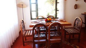 Un coin repas avec une table en bois, quatre chaises, un panier de fruits et une fenêtre donnant sur un paysage verdoyant.