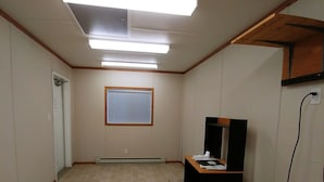 A room with a tiled floor, a desk with a computer and papers, a window with blinds, a door, and a ceiling light fixture. - Sundown Motel (Watrous)