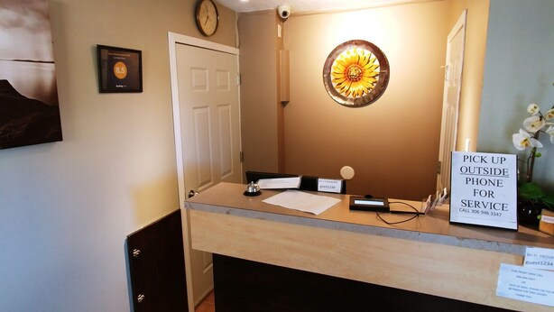A reception area with a desk, a clock, a framed certificate, a flower wall art, and a sign for pick-up outside phone service. - Sundown Motel (Watrous)
