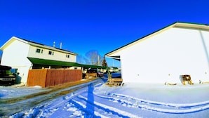 A snow-covered street with two white buildings, one with a green roof and another with a white roof. - Sundown Motel (Watrous)