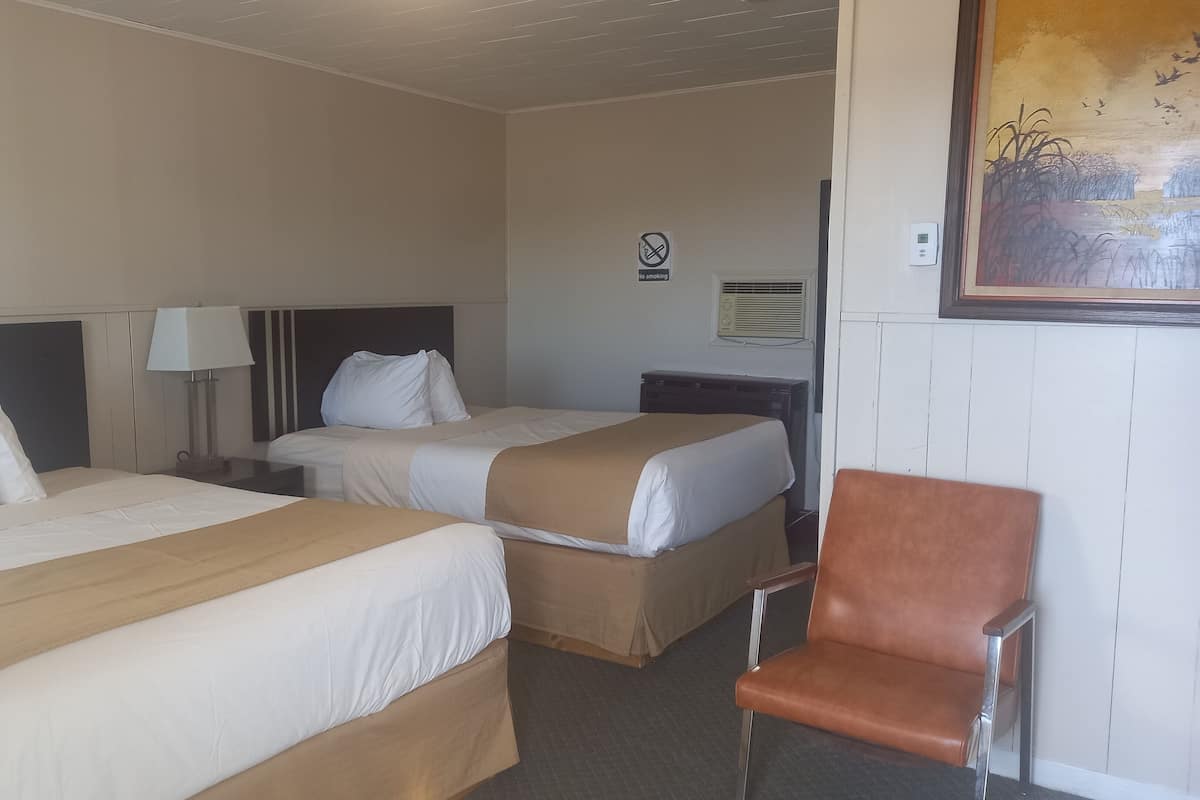Une chambre d’hôtel avec deux lits, une chaise, une table de chevet avec une lampe, un climatiseur fixé au mur et un panneau indiquant qu’il est interdit de fumer.