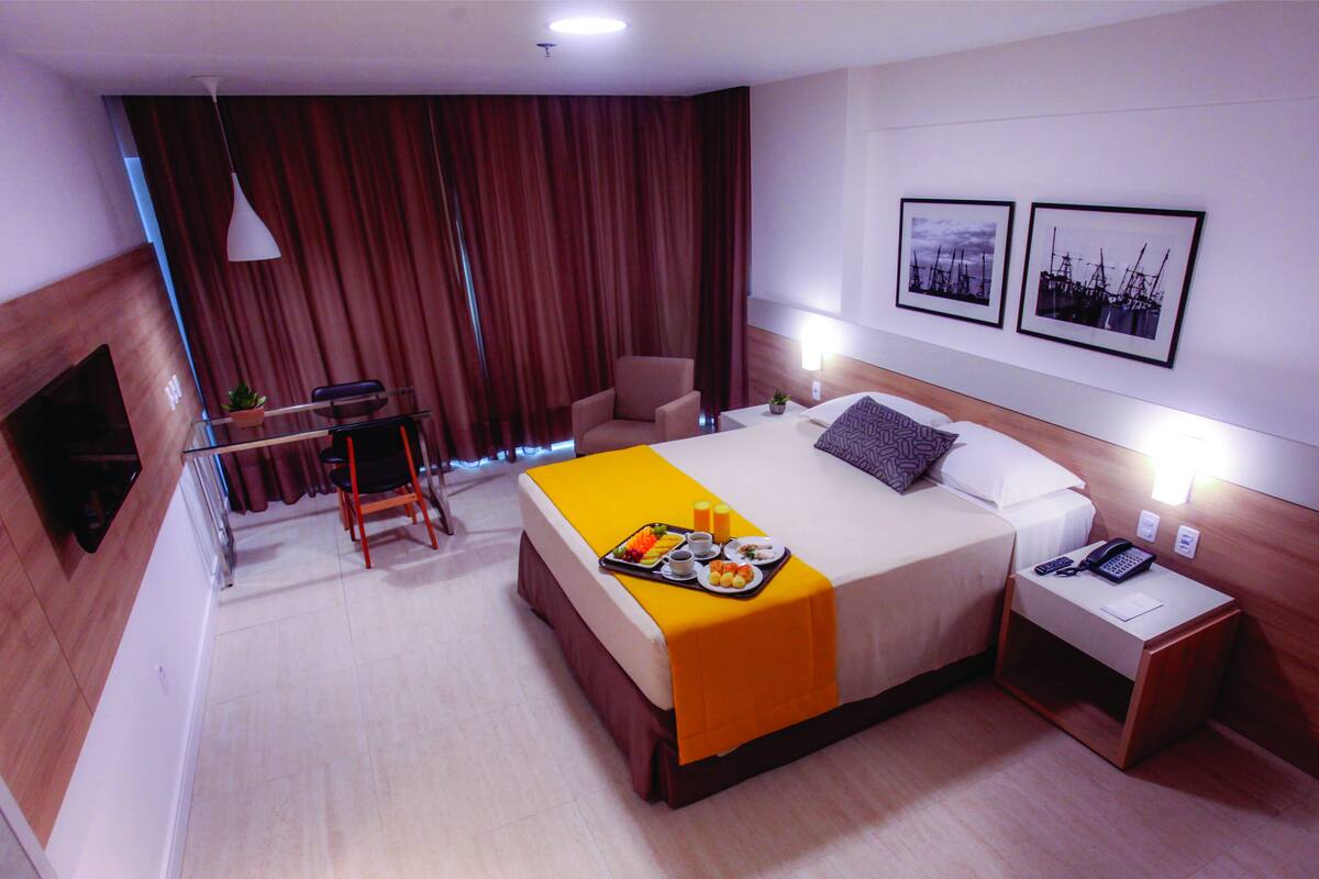A hotel room with a bed, bedside table, chair, and a desk with a plant.