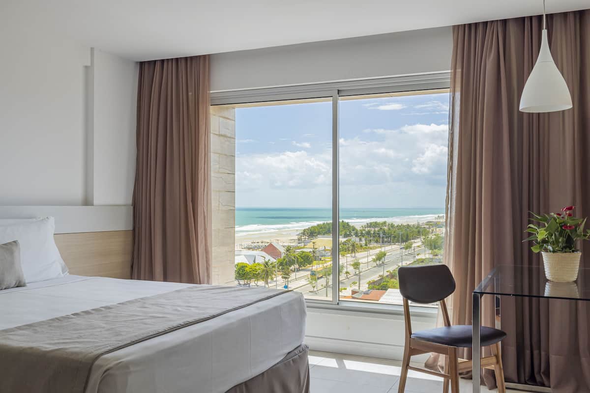 A hotel room with a large window offering a view of the beach and ocean.