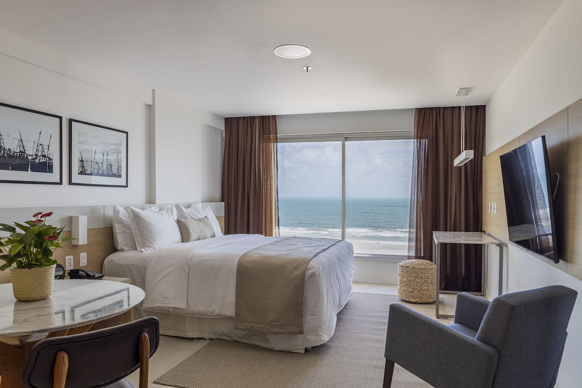 A hotel room with a bed, a desk, a chair, a TV, and a view of the ocean.