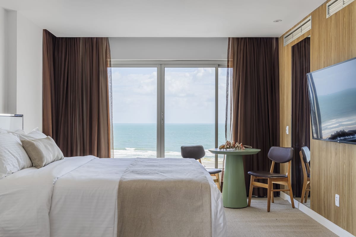 A hotel room with a large bed, a small table, a chair, a TV, and a view of the ocean.