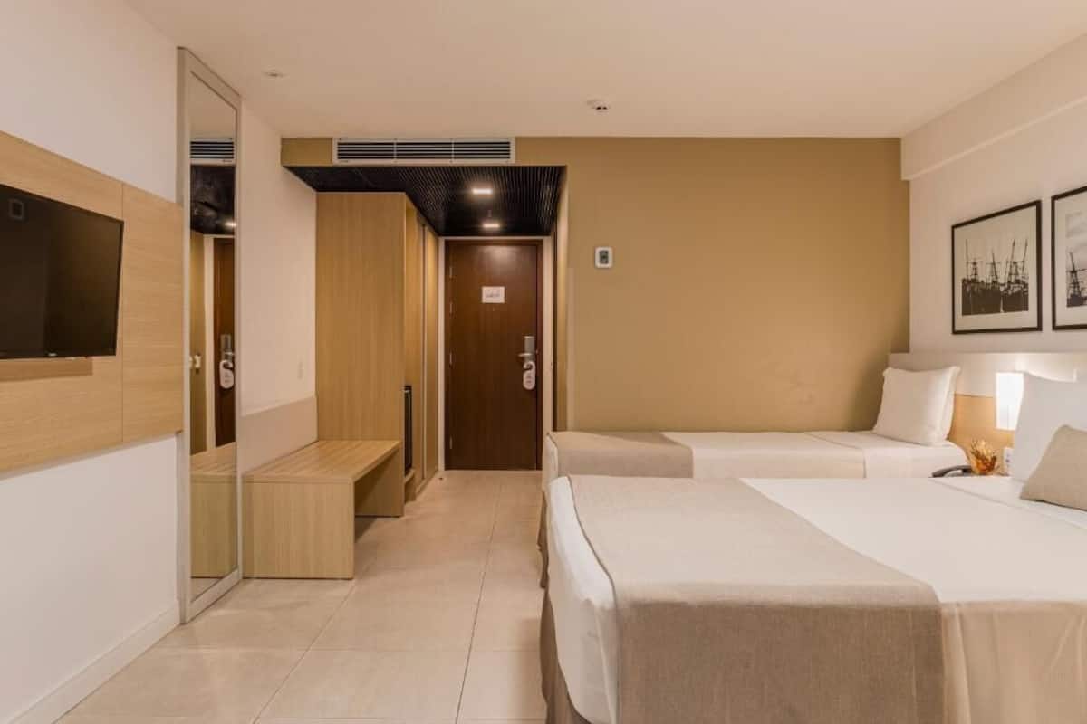 A hotel room with two beds, a wardrobe, a bench, and a television.