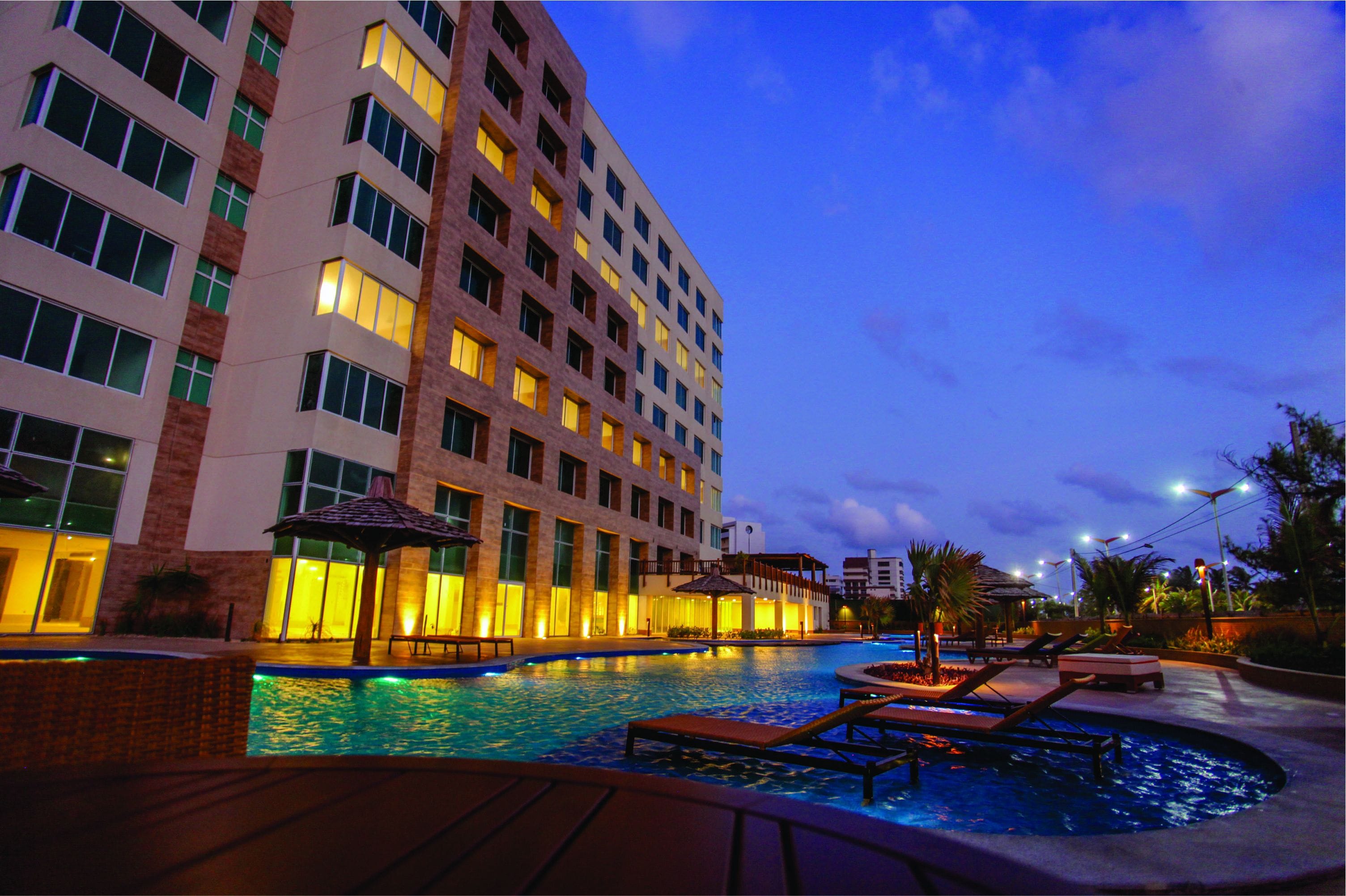 A hotel with a swimming pool, lounge chairs, and a well-lit building facade.