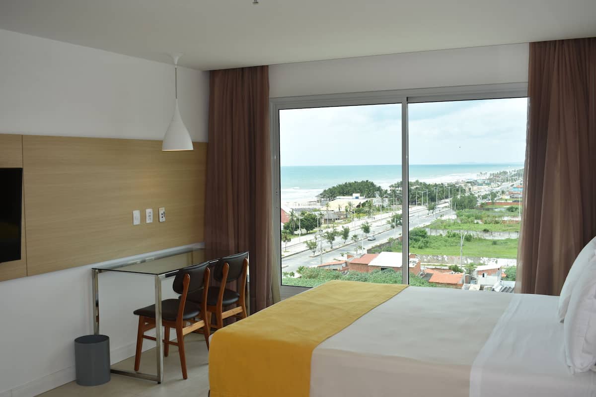 A hotel room with a bed, a desk with a chair, a TV, and a large window with a view of the beach and city.