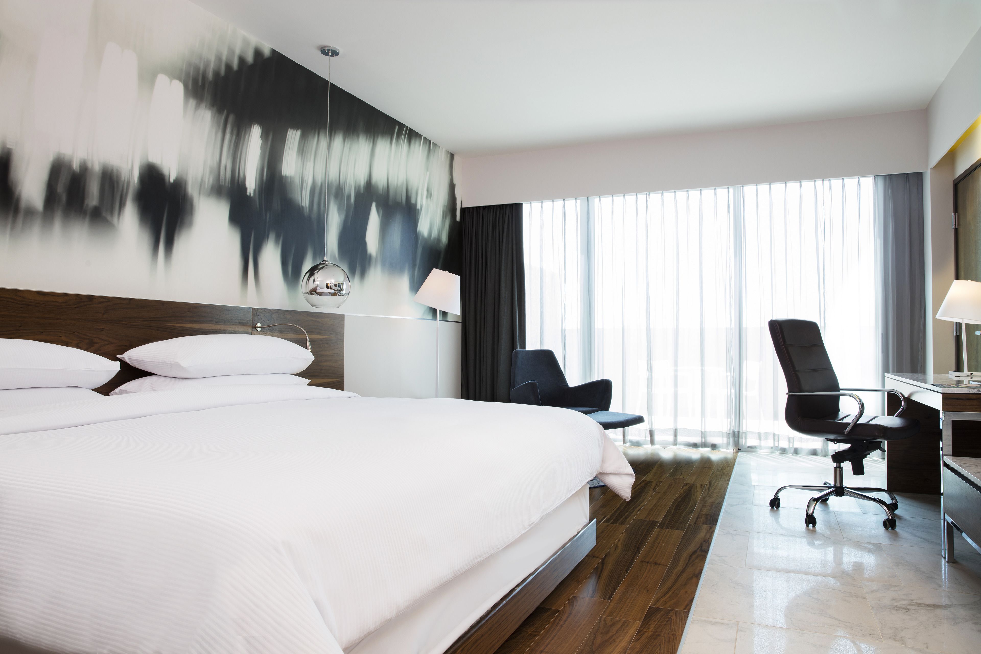 Ein modernes Hotelzimmer mit einem großen Bett, einem Schreibtisch mit Stuhl und einem großen Fenster mit Vorhängen.