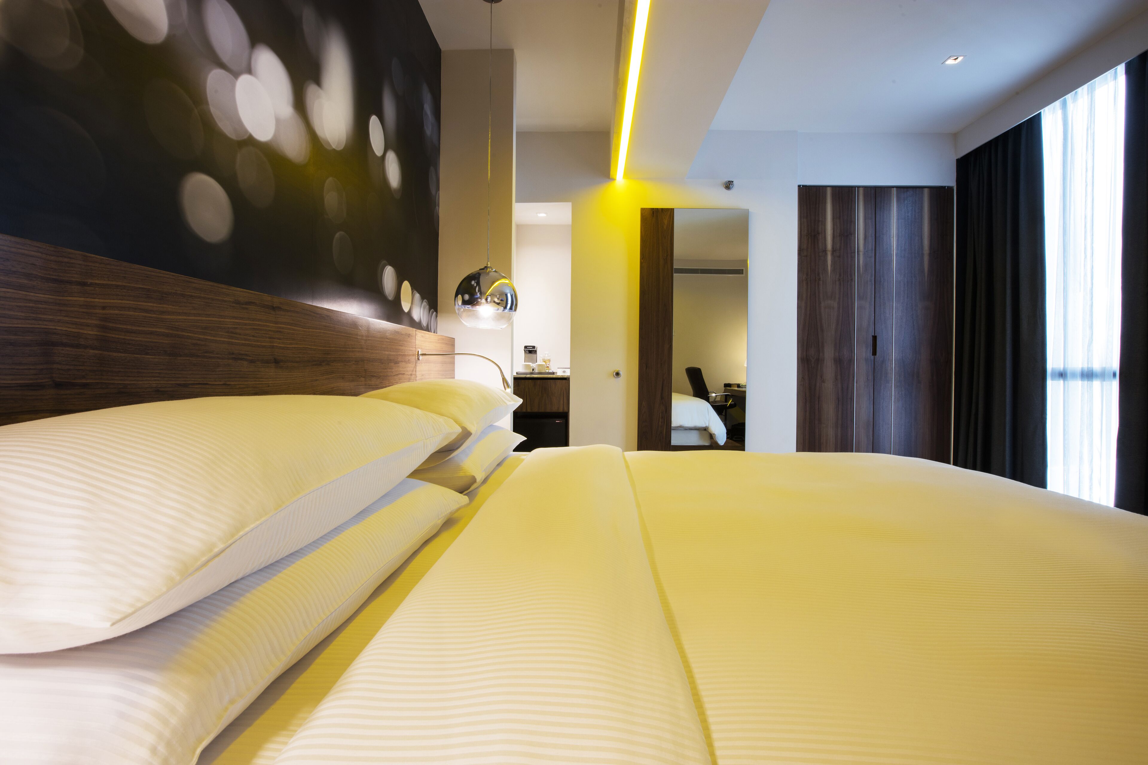 A modern hotel room with a large bed, a wooden headboard, a mirror, and a view of the city through large windows.