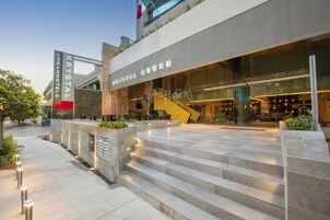 Una entrada de hotel moderna con fachada de vidrio, asientos al aire libre y una escalera que lleva a la entrada.