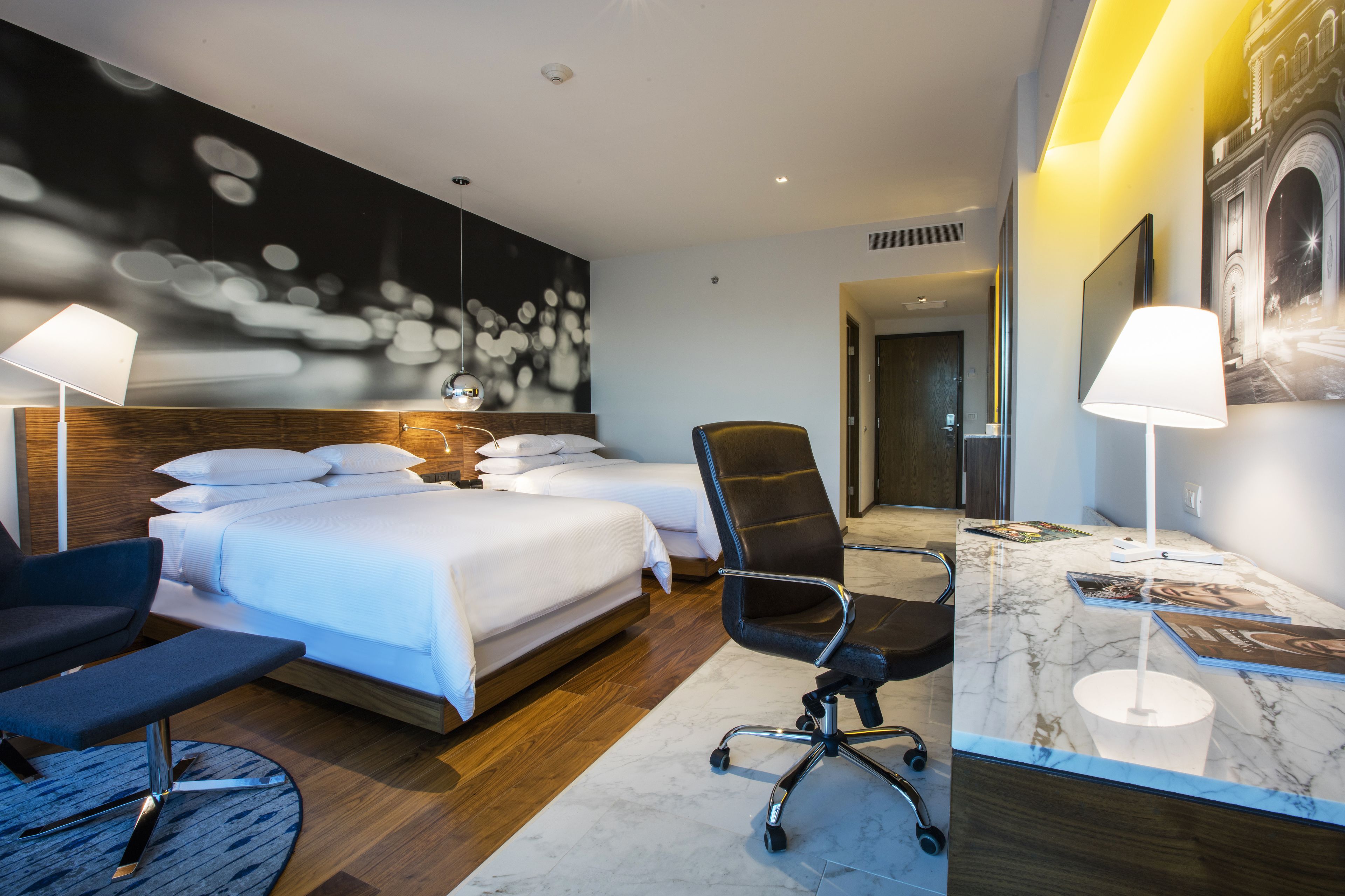 A modern hotel room with a large bed, two bedside tables, a desk with a chair, and a large wall mural.