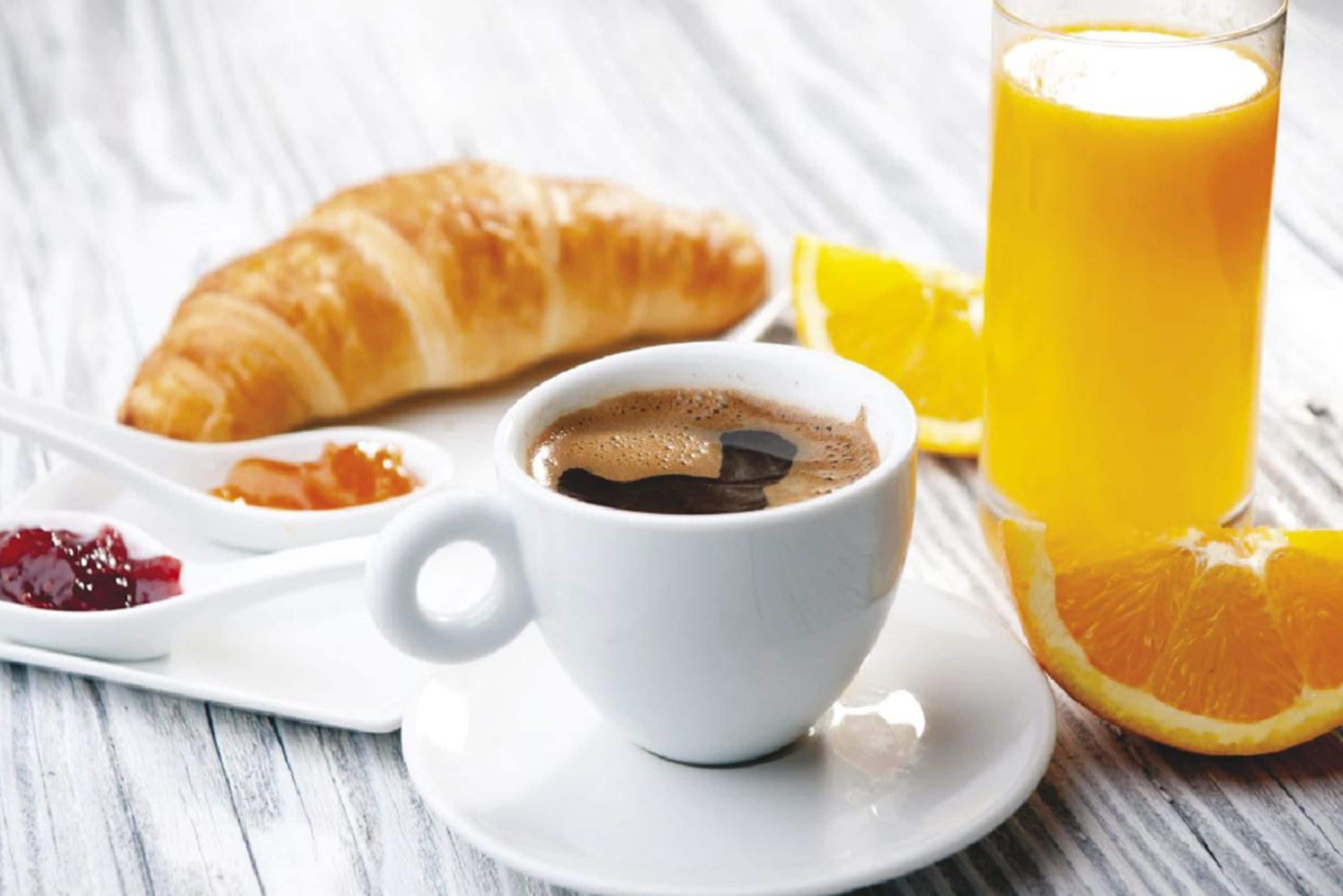Une table dressée pour le petit-déjeuner avec un croissant, une tasse de café, un verre de jus d’orange et une tranche d’orange sur une assiette blanche.