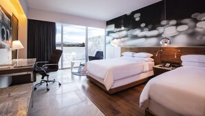 A modern hotel room with a large bed, a desk, and a chair. There is a view of the city through large windows. - Krystal Urban Guadalajara (Guadalajara)