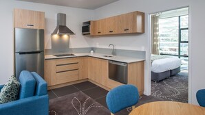 A modern kitchen with wooden cabinets, a stainless steel refrigerator, and a dishwasher. There is a dining area with a table and chairs, and a bedroom with a bed and a window. - One88 On Commerce (Whakatane)