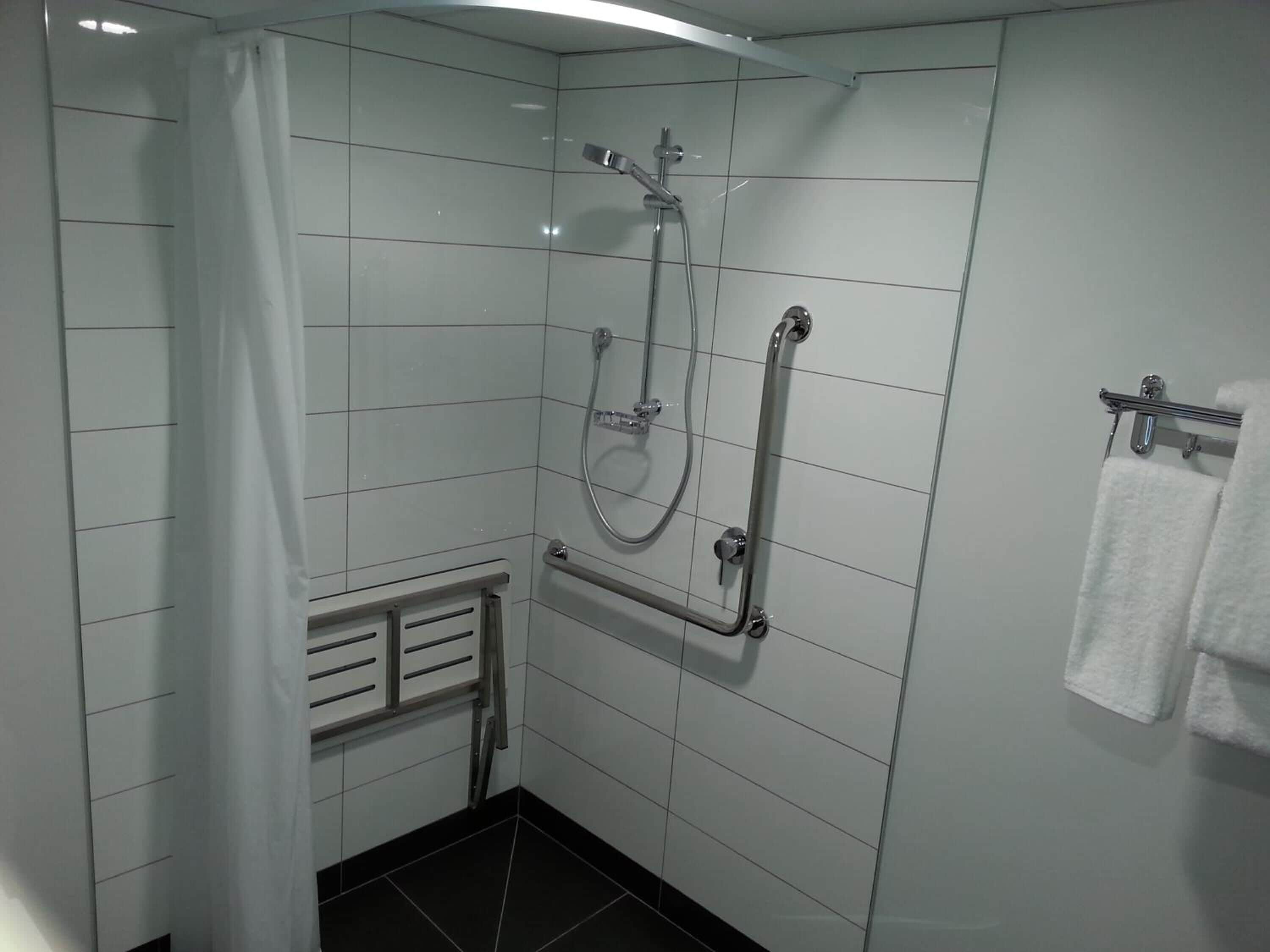 A bathroom with white tiled walls, a shower with a handheld showerhead, a towel rack with two towels, and a radiator.