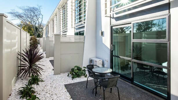 A modern building with a glass facade, a patio with a table and chairs, and a small garden area with plants. - One88 On Commerce (Whakatane)
