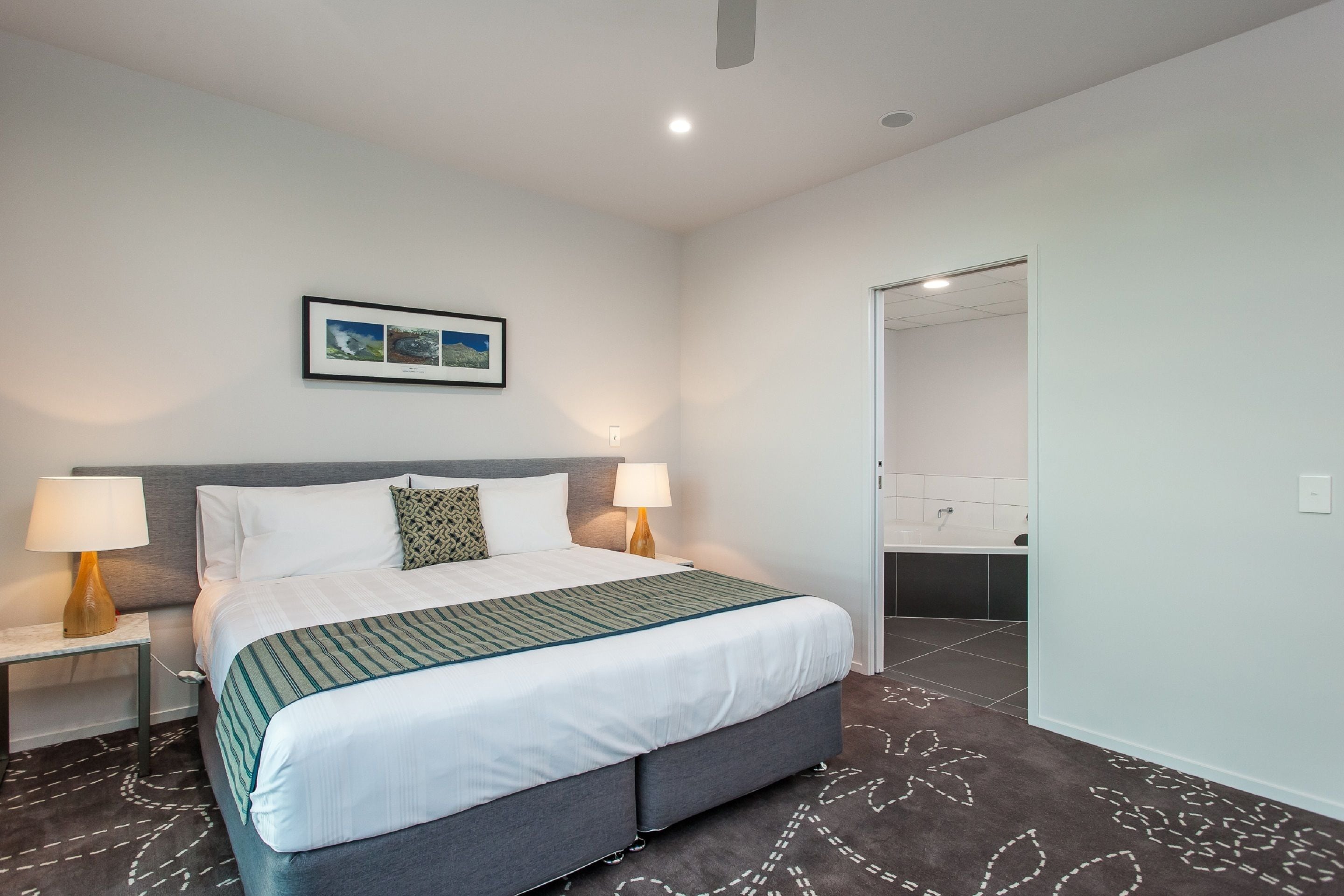 Una habitación de hotel moderna con una cama grande, dos lámparas de noche, un cuadro enmarcado en la pared y un baño visible a través de una puerta abierta.