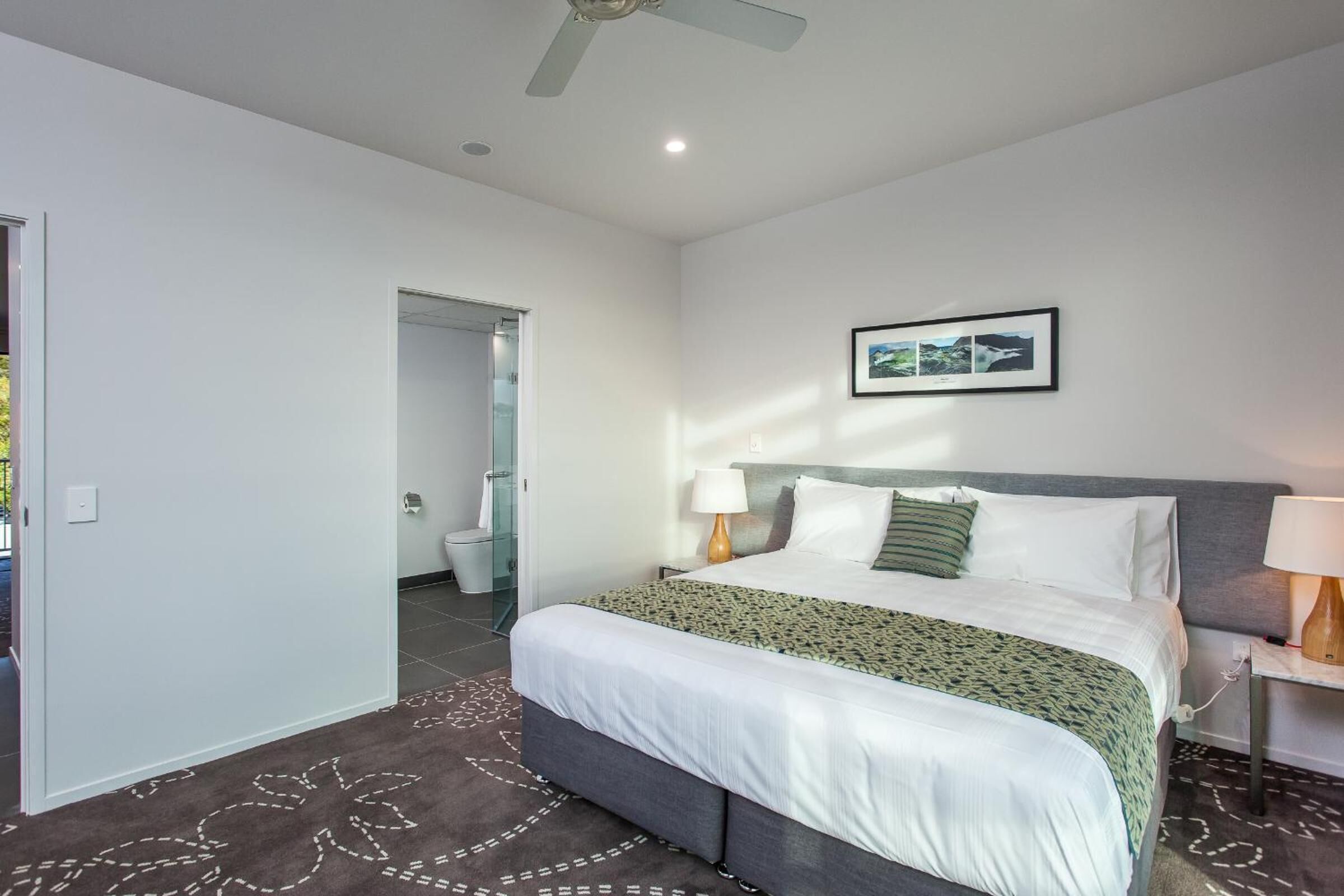 A modern hotel room with a large bed, a ceiling fan, a framed picture on the wall, and a bathroom visible through an open door.