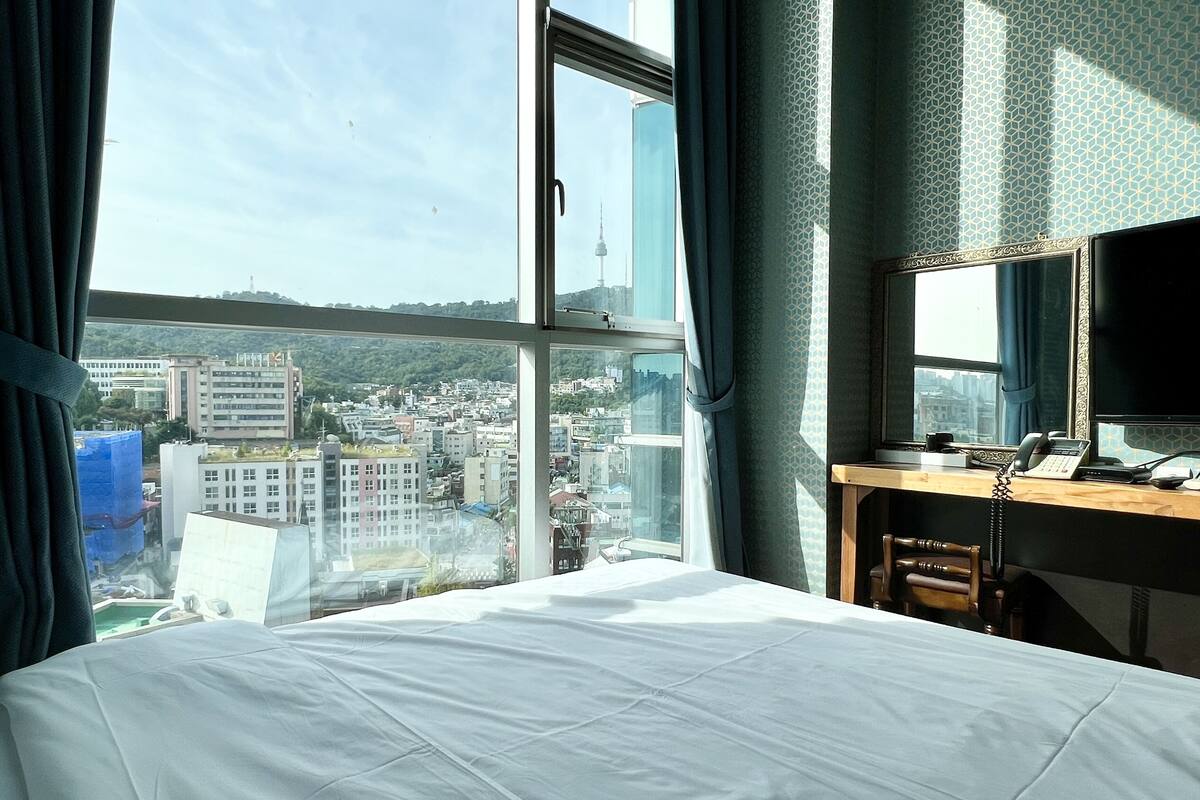 A hotel room with a large window, a bed, a desk with a telephone, and a mirror.