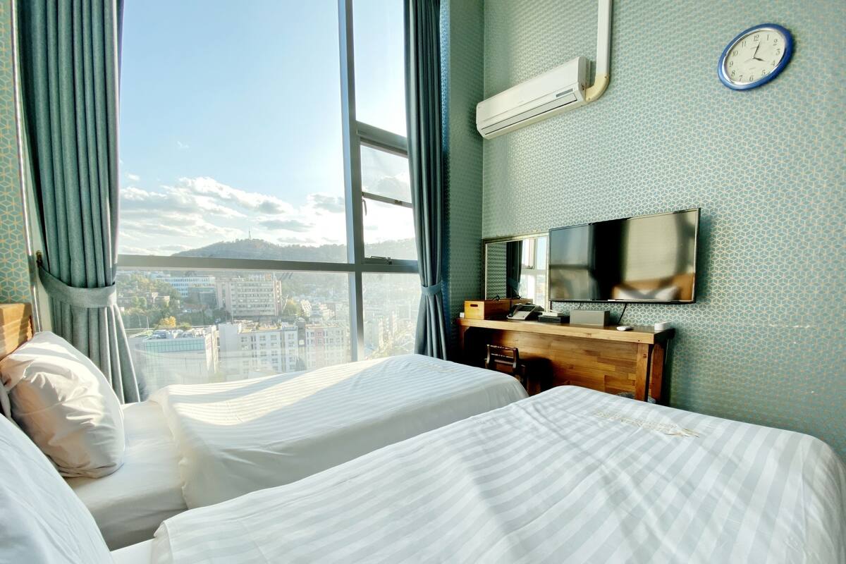 A hotel room with two beds, a large window with a view of a cityscape, an air conditioning unit, a clock, and a desk with a mirror and a flat-screen TV.