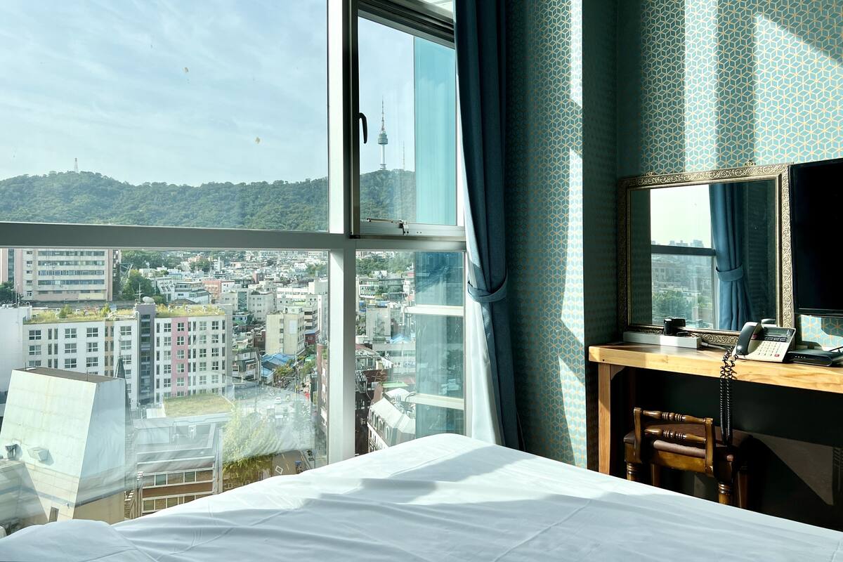 A hotel room with a large window offering a city view, a desk with a telephone, and a chair.
