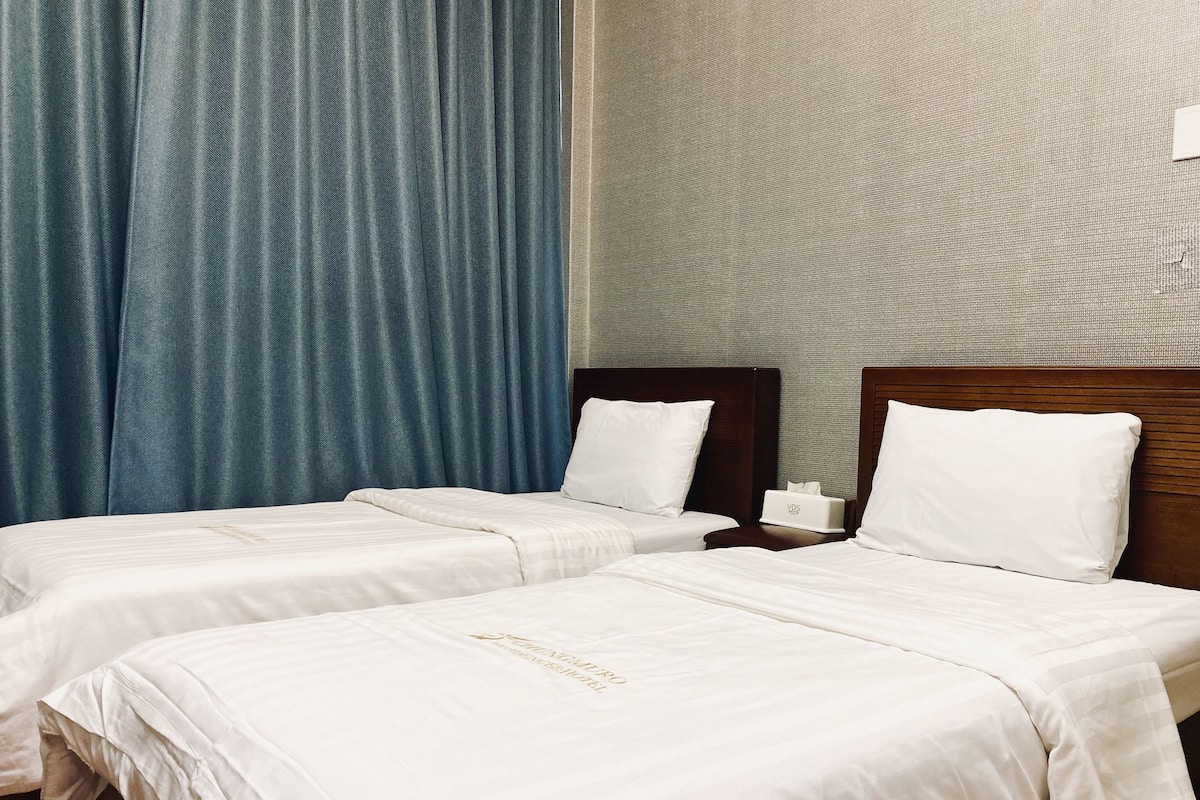 A hotel room with two beds, a wooden headboard, a nightstand with a tissue box, and a blue curtain.