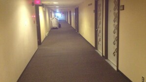 A hotel hallway with carpeted floors, doors on the right, and a lighted exit sign on the left. - Classic Inn (Melville)