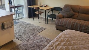 Une pièce confortable avec un lit, un canapé, un bureau et une chaise. Il y a une fenêtre avec des rideaux, un tableau encadré au mur et une petite table sur laquelle se trouvent une bouteille et des en-cas.