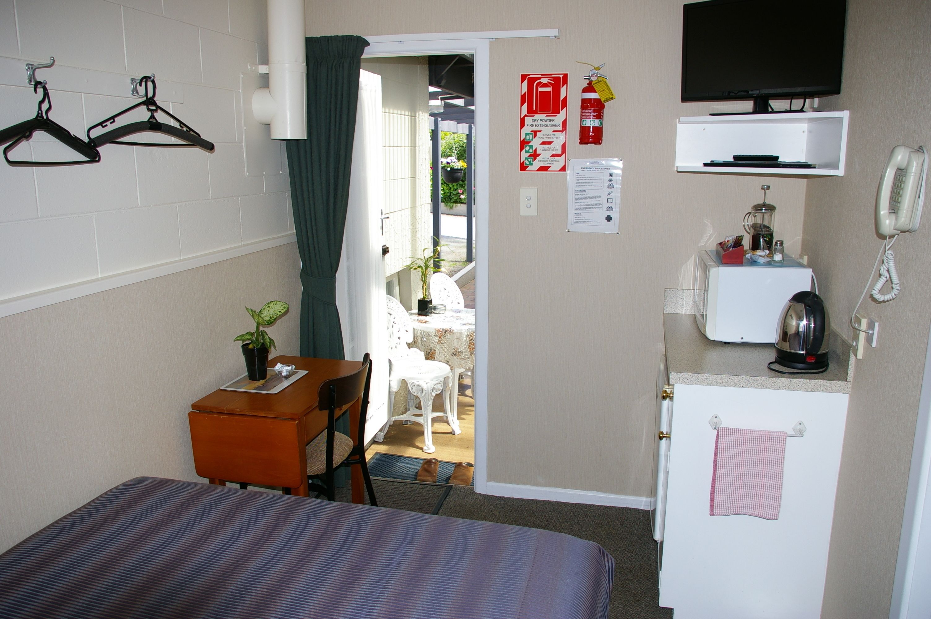 Une chambre d’hôtel compacte comprenant un lit, un petit bureau avec une chaise, une kitchenette équipée d’un réfrigérateur, une télévision et une porte donnant sur un balcon.