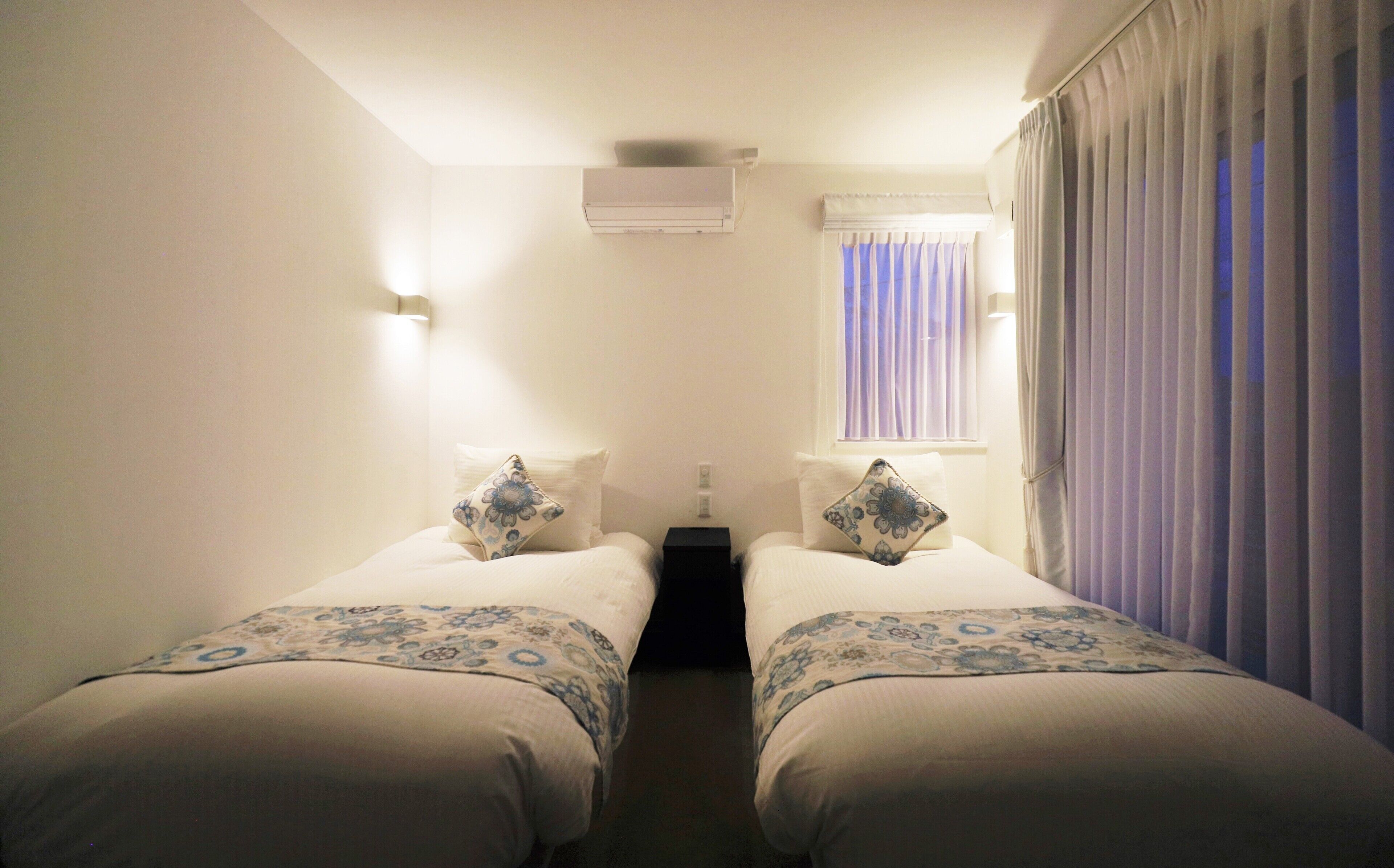 Habitación doble con dos camas, una ventana con cortinas, un equipo de aire acondicionado y una mesita de noche.