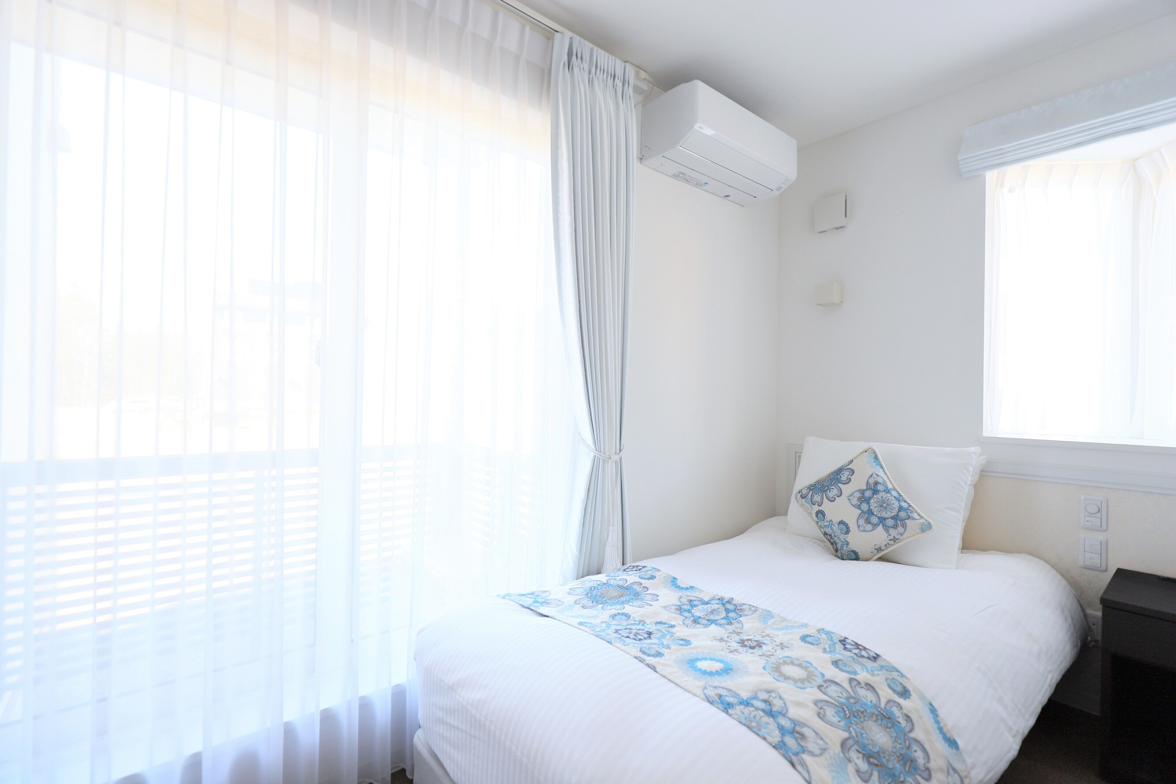 A neatly made bed with a floral-patterned bedspread, a bedside table, and a window with sheer curtains.