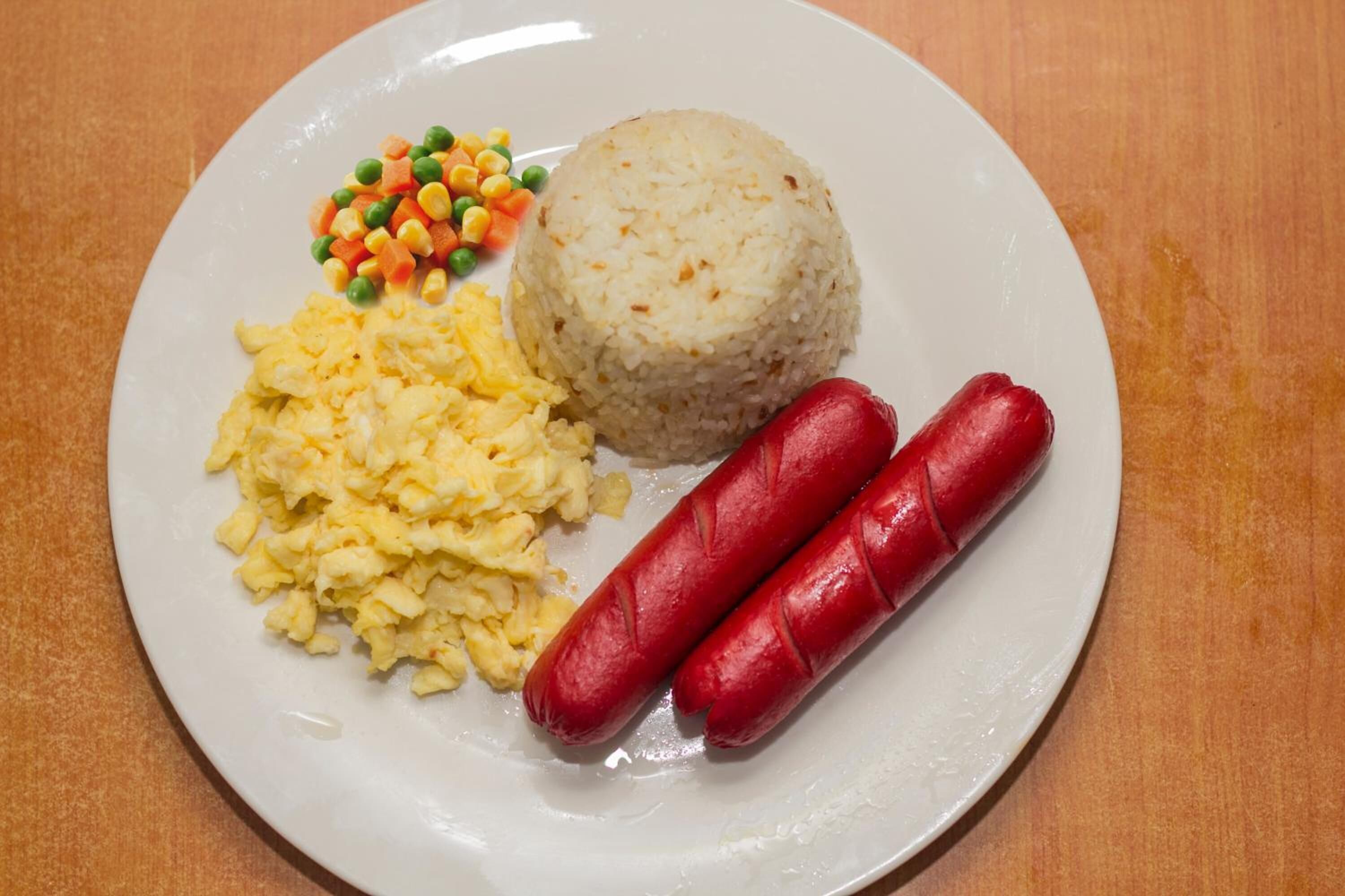Une assiette contenant des œufs brouillés, une portion de riz, deux saucisses et un accompagnement de légumes variés.