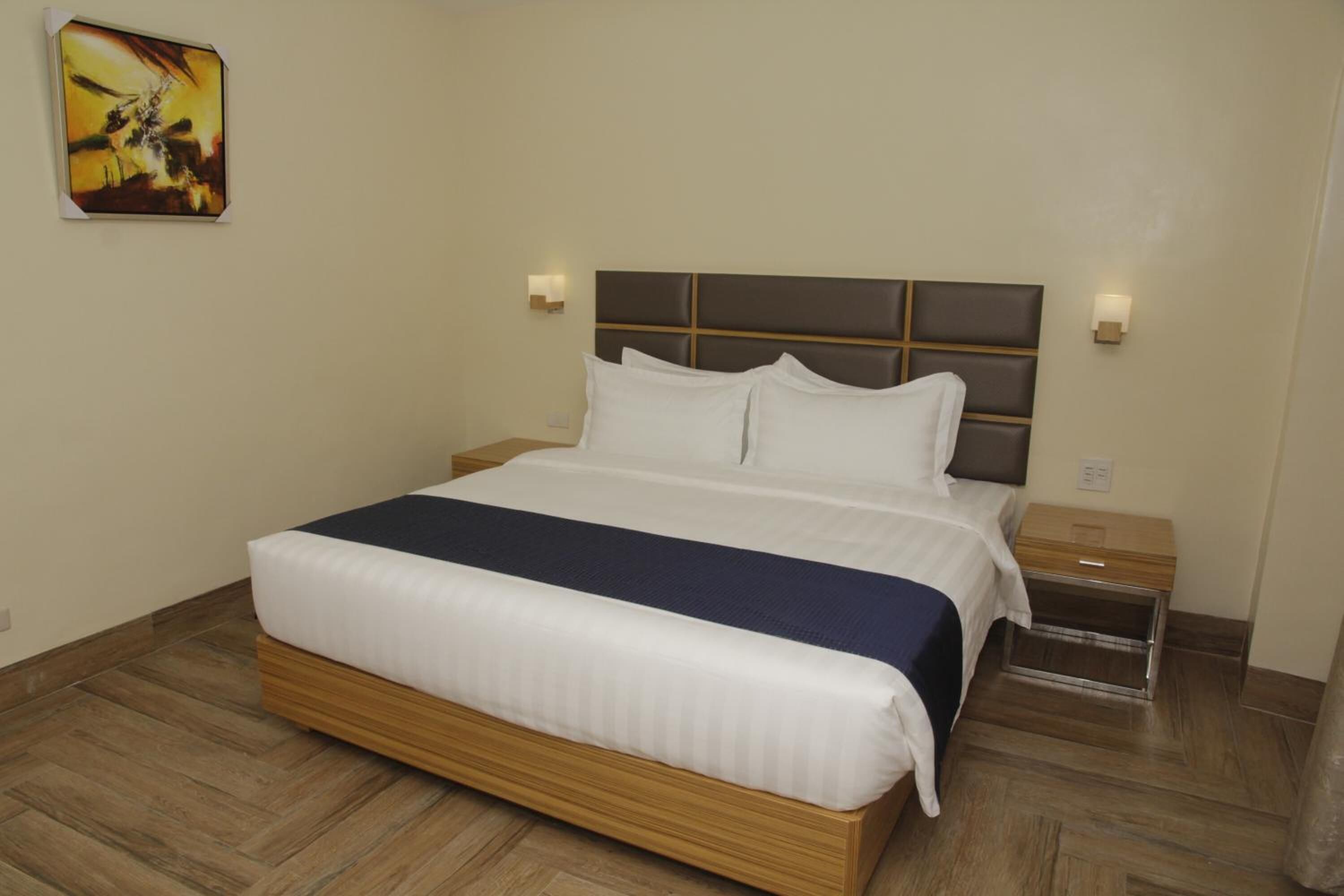 Quarto de hotel com uma cama grande, cabeceira de madeira, duas mesas de cabeceira e um criado-mudo.