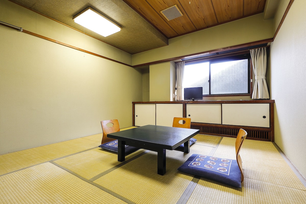 Um espaço com tatames, uma mesa baixa, duas almofadas, uma janela com cortinas e uma televisão sobre um móvel.