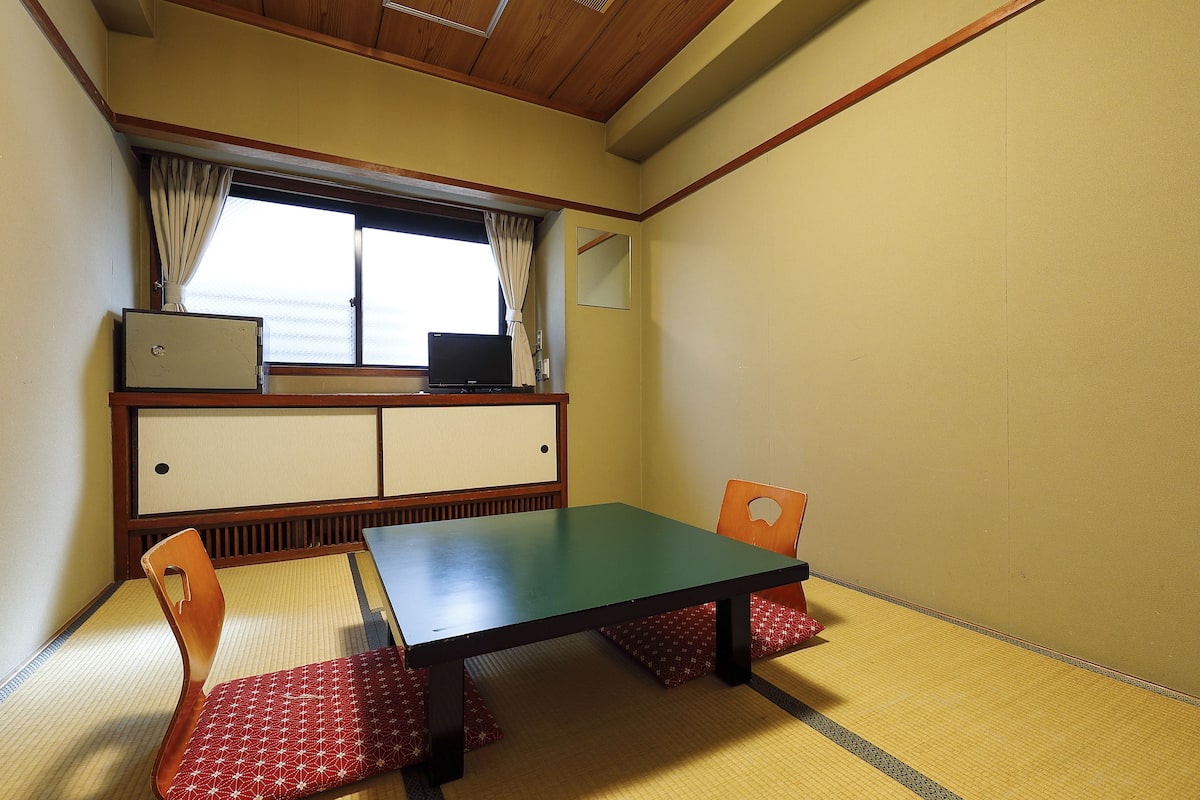 Um quarto japonês tradicional com tatames, uma mesa baixa, duas cadeiras, um futon e uma televisão, além de uma janela com cortinas.