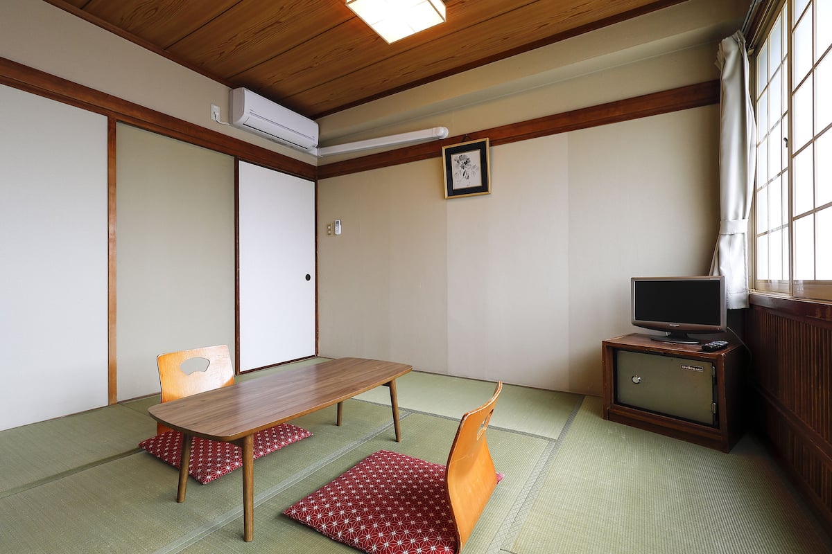 Um quarto japonês tradicional com piso de tatame, uma mesa baixa, duas cadeiras, uma televisão em um móvel e uma porta deslizante.