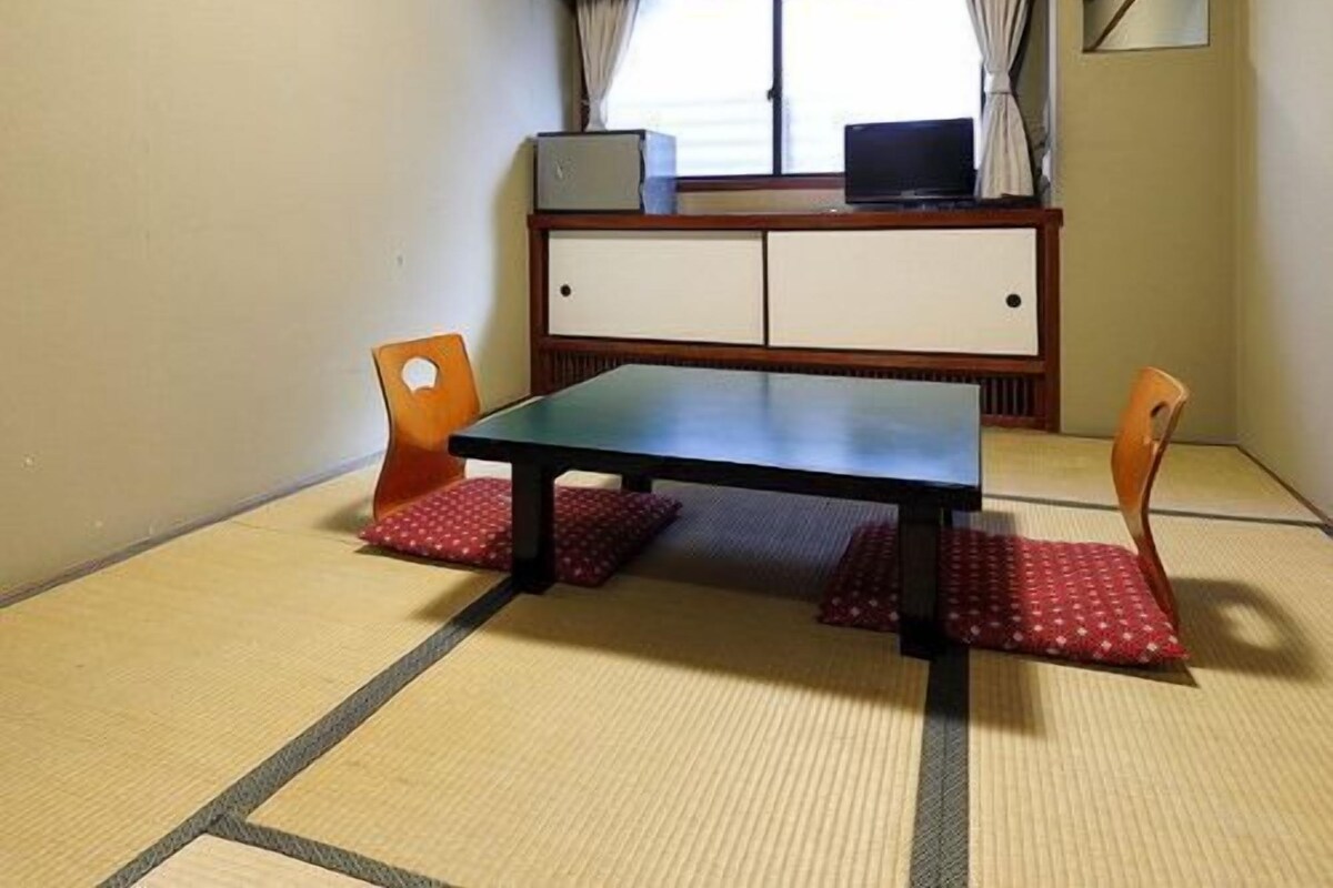 Una habitación con suelo de tatami, una mesita baja, dos sillas, un mueble bajo y una ventana con cortinas.