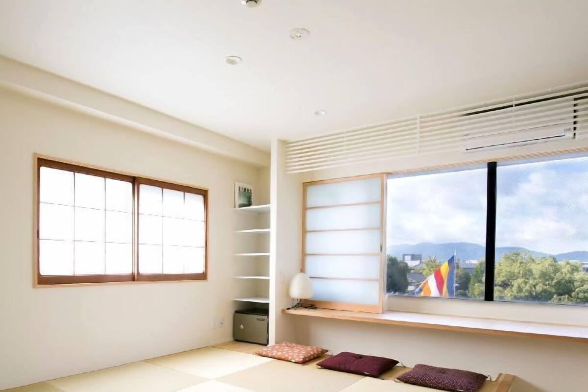 Quarto com piso de tatame, um biombô shoji, uma pequena prateleira e uma janela com vista para montanhas.